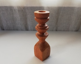 May include: A wooden candle holder with a natural finish. The holder has a square base and a cylindrical top with a rounded opening for a candle.