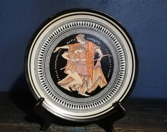 Może przedstawiać: Czarno-biały talerz ceramiczny z namalowaną sceną przedstawiającą dwie kobiety w pomarańczowych i różowych szatach. Talerz ma czarno-białą obwódkę z greckim wzorem klucza i tekst "GOTIS PHASCI ENAQE NOAOTZAMOLH OINOF LEADOS" wokół krawędzi.