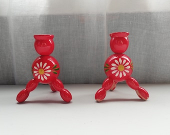 May include: Two red wooden candle holders with a daisy design. Each candle holder has a round base with three legs, a spherical middle section with a painted daisy, and a top cup for a candle. The candle holders are painted red.