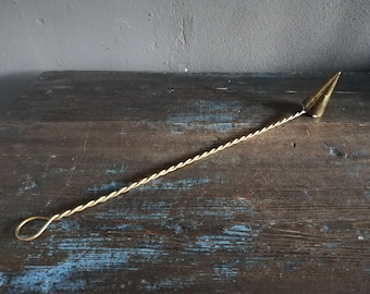 May include: A gold-colored metal candle snuffer with a twisted handle and a pointed tip. The snuffer is lying on a dark wooden surface.