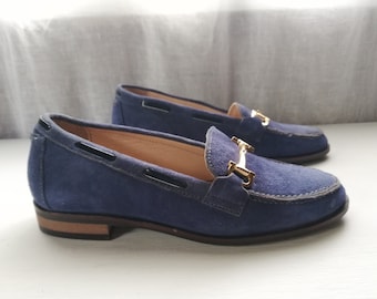 May include: A pair of blue suede loafers with gold buckles. The loafers have a pointed toe and a low heel.