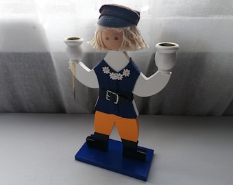Puede incluir: Un portavelas de madera con forma de persona que lleva un atuendo azul y blanco, un sombrero marrón y sostiene dos vasos de vela blancos.