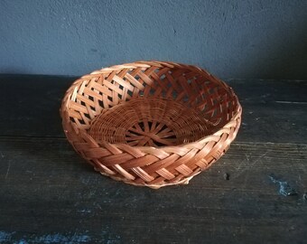 May include: A round, woven basket made of natural brown wicker. The basket has a wide, open top and a slightly rounded bottom. It is perfect for holding small items or displaying decorative objects.