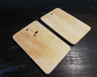 May include: Two wooden cutting boards with a light brown color. Both boards have a hole in the top for hanging. One board has a dark brown burn mark.