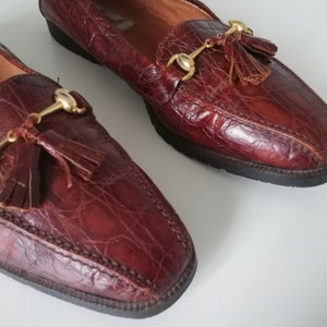 Puede incluir: Un par de mocasines de cuero marr&oacute;n con borlas doradas y una hebilla de metal dorado. Los mocasines tienen un patr&oacute;n de piel de cocodrilo.