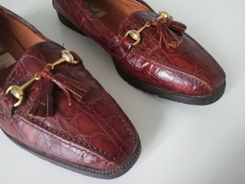 Puede incluir: Un par de mocasines de cuero marr&oacute;n con borlas doradas y una hebilla de metal dorado. Los mocasines tienen un patr&oacute;n de piel de cocodrilo.