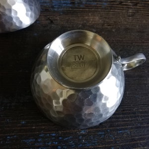 May include: A silver hammered metal cup with a handle. The bottom of the cup has the text "TW R10" stamped on it.