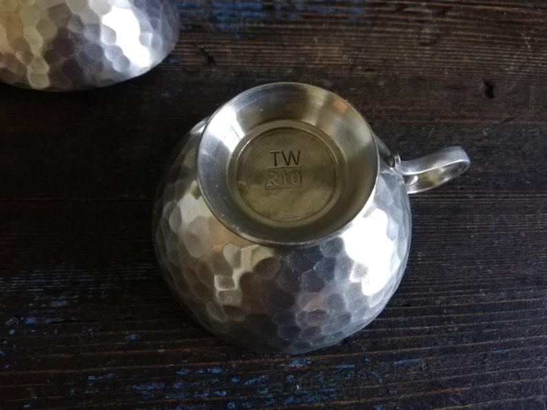 May include: A silver hammered metal cup with a handle. The bottom of the cup has the text "TW R10" stamped on it.