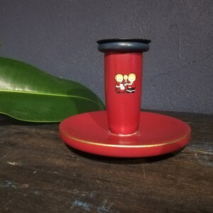 May include: Red candle holder with a gold rim and a black top. The holder features a design of two children in red and white clothing.