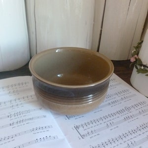 May include: A ceramic bowl with a brown and tan color scheme. The bowl has a dark brown band around the middle and is sitting on a sheet of musical notes. The background includes white cylindrical objects.