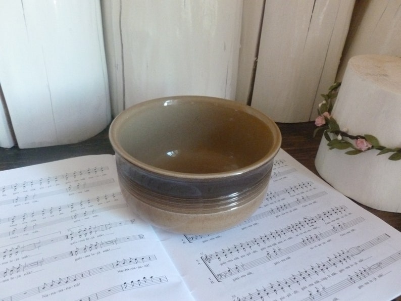 May include: A ceramic bowl with a brown and tan color scheme. The bowl has a dark brown band around the middle and is sitting on a sheet of musical notes. The background includes white cylindrical objects.
