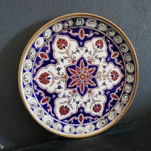 May include: A decorative ceramic plate with a white background and intricate blue, red, and gold floral patterns. The plate has a gold rim and is perfect for adding a touch of elegance to any home.