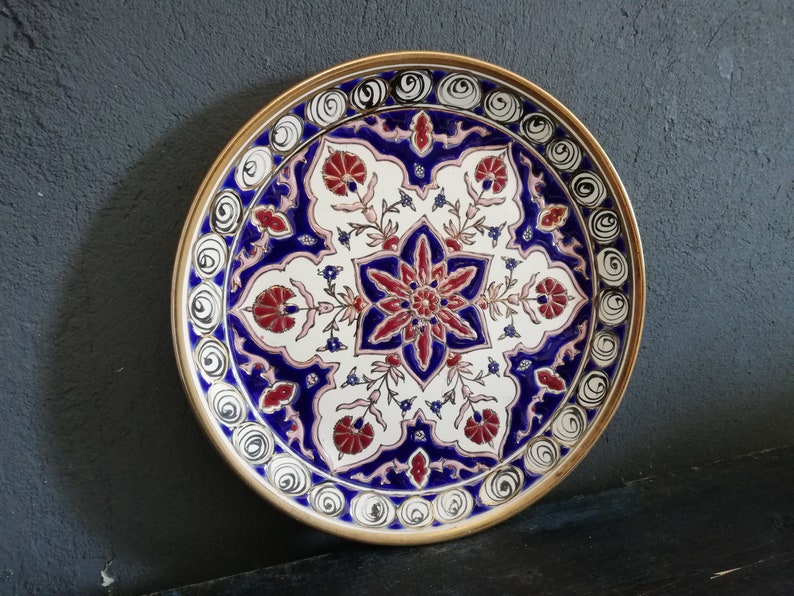 May include: A decorative ceramic plate with a white background and intricate blue, red, and gold floral patterns. The plate has a gold rim and is perfect for adding a touch of elegance to any home.