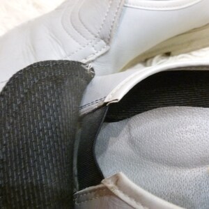 May include: A pair of gray leather shoes with black elastic straps and a padded insole. The shoes have a textured surface and a simple design.