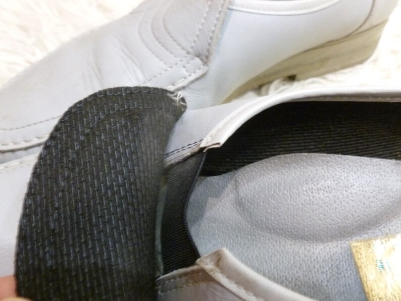 May include: A pair of gray leather shoes with black elastic straps and a padded insole. The shoes have a textured surface and a simple design.