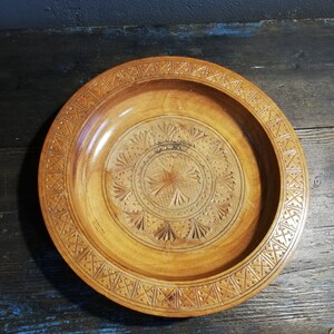 May include: A round wooden serving bowl with intricate carvings. The bowl has a light brown color and a geometric pattern around the rim.