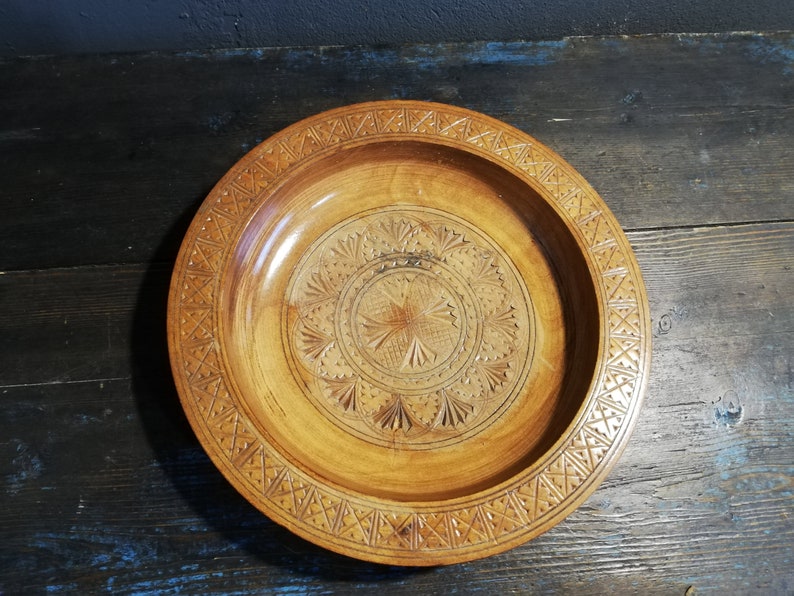 May include: A round wooden serving bowl with intricate carvings. The bowl has a light brown color and a geometric pattern around the rim.