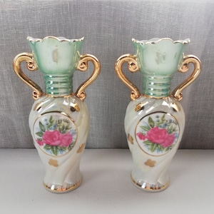 May include: Two white porcelain vases with gold accents and floral designs. The vases have a green and white swirl pattern and feature a pink rose with green leaves in a circular design.