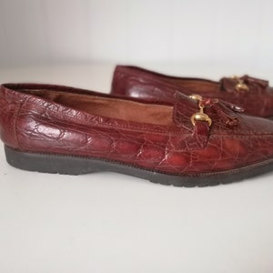 Puede incluir: Un par de mocasines de cuero marr&oacute;n con un detalle de borla y detalles dorados. Los mocasines tienen un patr&oacute;n de piel de cocodrilo.