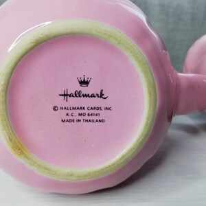 May include: A pink ceramic mug with a scalloped rim and a handle. The bottom of the mug has a black and white Hallmark logo with the text "Hallmark Cards, Inc. K.C., MO 64141 Made in Thailand."