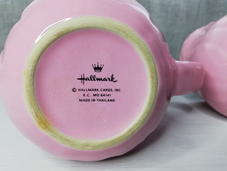 May include: A pink ceramic mug with a scalloped rim and a handle. The bottom of the mug has a black and white Hallmark logo with the text "Hallmark Cards, Inc. K.C., MO 64141 Made in Thailand."