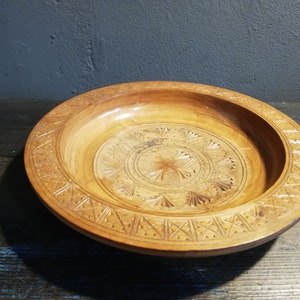 May include: A wooden bowl with intricate carvings in a circular pattern. The bowl is light brown and has a smooth, polished finish.