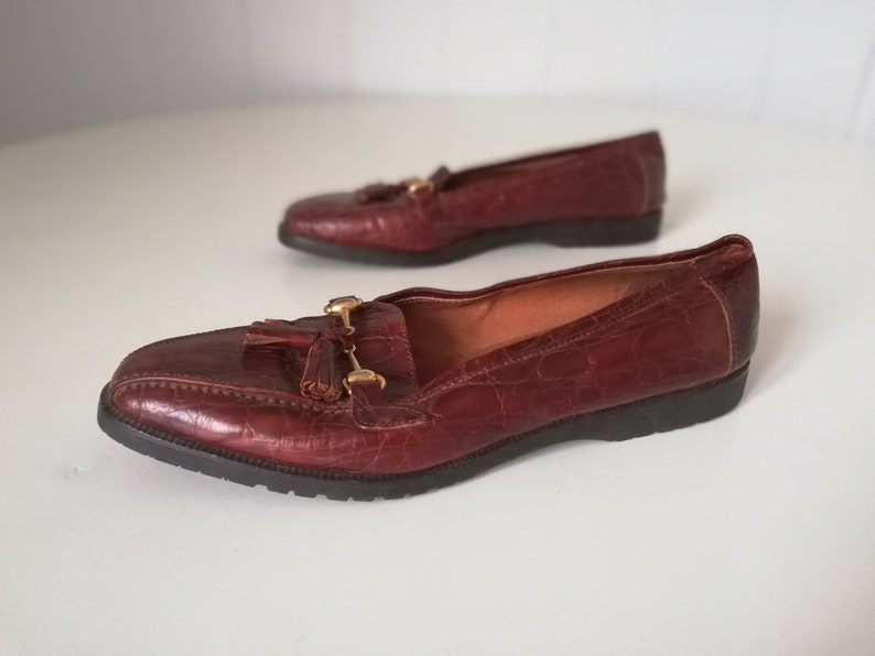 Puede incluir: Un par de mocasines de cuero marr&oacute;n con detalles dorados y borlas. Los mocasines tienen una parte superior de cuero texturizado y una suela de goma negra.