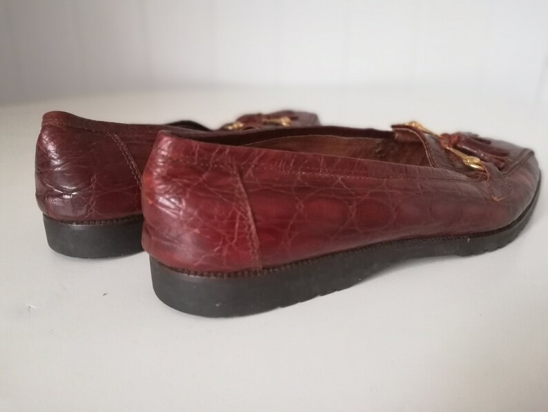 Puede incluir: Un par de mocasines de cuero marr&oacute;n con un detalle de borla y herrajes dorados. Los mocasines tienen un patr&oacute;n texturizado, similar al de un cocodrilo.
