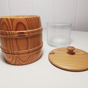 Puede incluir: Un dispensador de azúcar de madera con forma de barril con tapa y un tarro de vidrio en el interior. La tapa tiene un pomo de madera y una pequeña ranura para sacar azúcar.
