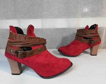 May include: A pair of red suede ankle boots with brown leather straps and buckles. The boots have a block heel and a pointed toe.