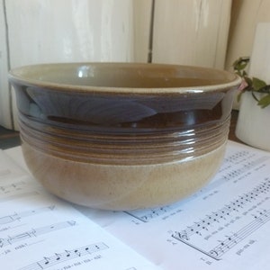 May include: A ceramic bowl with a two-tone brown and tan glaze. The bowl has a rounded shape with a wide rim and horizontal textured bands around the middle. The bowl is sitting on a sheet of musical notes.