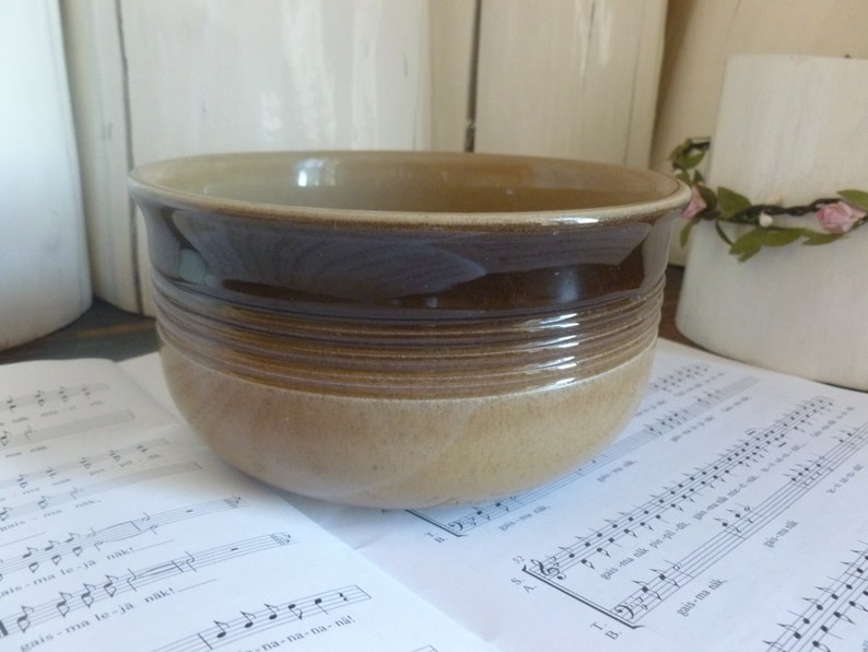 May include: A ceramic bowl with a two-tone brown and tan glaze. The bowl has a rounded shape with a wide rim and horizontal textured bands around the middle. The bowl is sitting on a sheet of musical notes.
