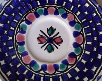 Könnte beinhalten: Eine weiße Keramik-Untertasse mit einem blauen und weißen geometrischen Muster am Rand. Die Mitte der Untertasse zeigt ein stilisiertes Blumenmuster in Rosa, Grün und Schwarz.