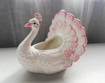 Puede incluir: Macetero de cerámica blanca en forma de pavo real con cola rosa. El macetero tiene forma de pavo real con un diseño detallado y una pequeña abertura para plantar.