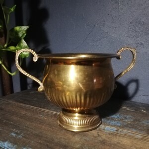 Pode incluir: Um vaso de metal dourado com duas alças ornamentadas. O vaso tem um design canelado e uma base redonda.