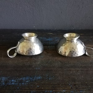 May include: Two silver hammered metal cups with handles. The cups are sitting on a dark wooden surface.