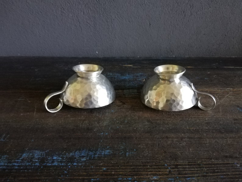 May include: Two silver hammered metal cups with handles. The cups are sitting on a dark wooden surface.