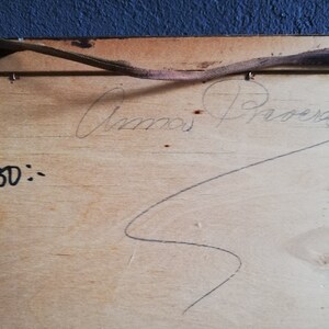May include: A close-up of the back of a wooden frame with a handwritten signature that reads "Anna Proera" and the number "50" written below. The frame has a brown leather strap attached to it.