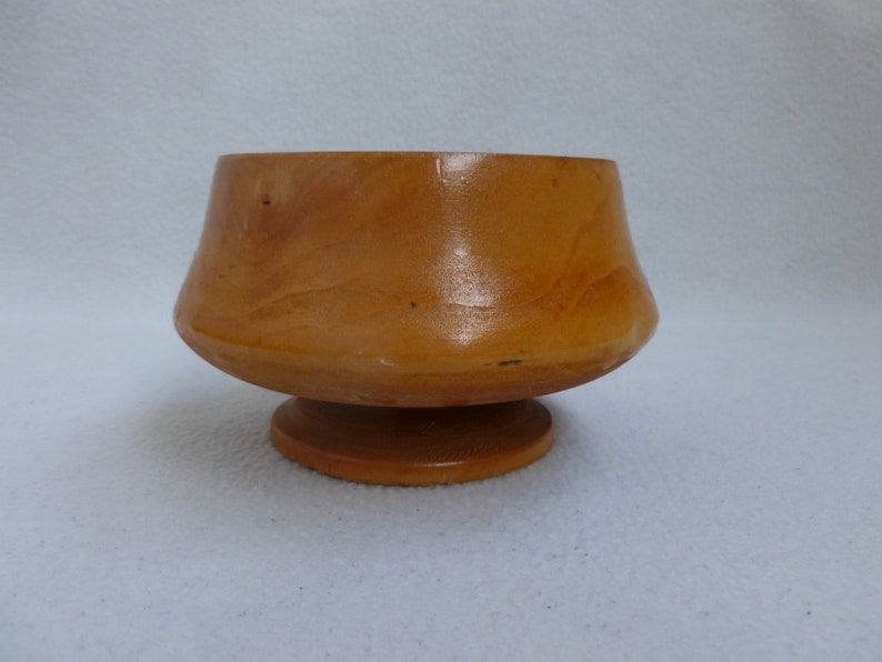 May include: A small, wooden bowl with a flared rim and a pedestal base. The bowl is made of light brown wood and has a smooth, polished finish.