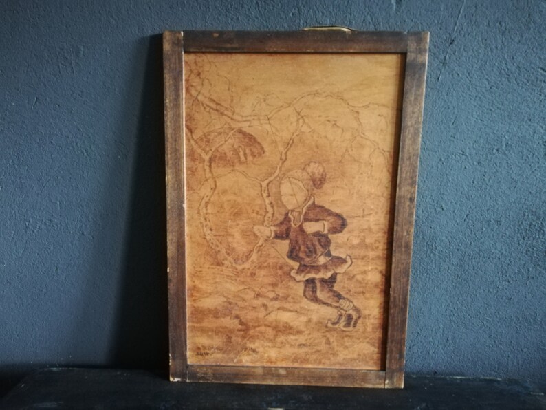May include: A framed wood-burned artwork depicting a girl in a winter scene. The girl is wearing a hat and coat and is holding a stick. The background is a forest with trees and snow.