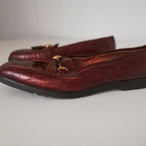 Puede incluir: Un par de mocasines de cuero marr&oacute;n con un detalle de borla y herrajes dorados. Los mocasines tienen una parte superior de cuero texturizado, con estampado de cocodrilo.