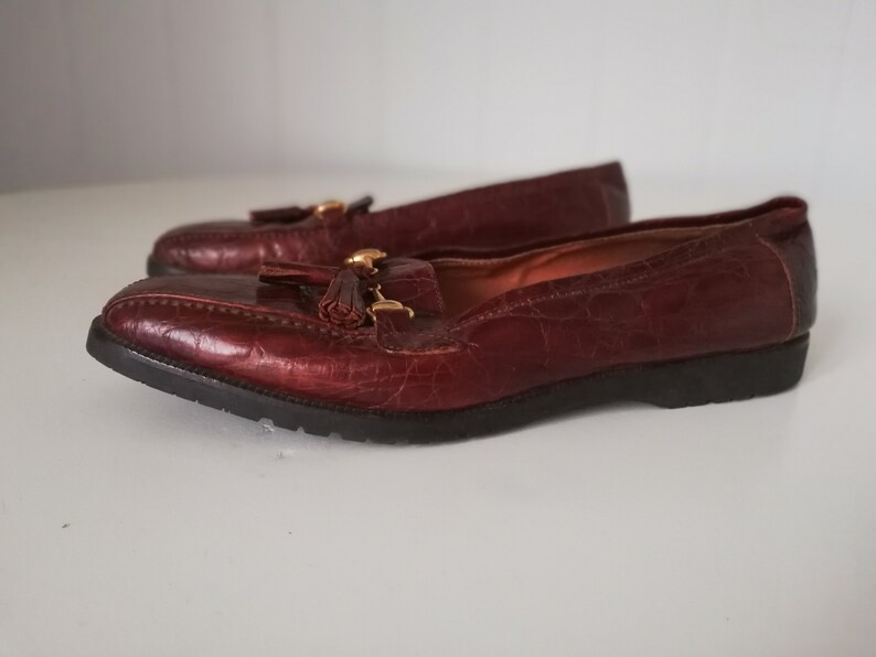 Puede incluir: Un par de mocasines de cuero marr&oacute;n con un detalle de borla y herrajes dorados. Los mocasines tienen una parte superior de cuero texturizado, con estampado de cocodrilo.