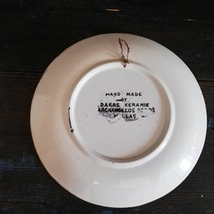 May include: White ceramic plate with a black hand-painted design. The back of the plate has the text "HAND MADE BY DAKAS KERAMIK ARCHANGELOS  8505  GLAS".