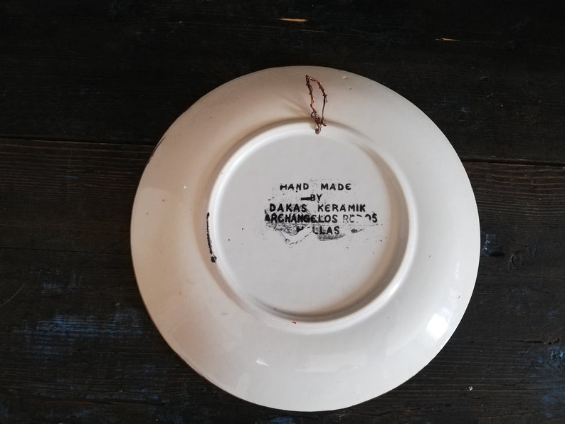 May include: White ceramic plate with a black hand-painted design. The back of the plate has the text "HAND MADE BY DAKAS KERAMIK ARCHANGELOS  8505  GLAS".