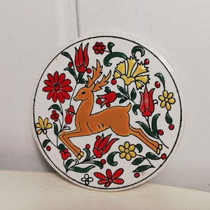 Puede incluir: Un posavasos de cer&aacute;mica blanca con un dise&ntilde;o floral colorido que presenta un ciervo en el centro. El ciervo es naranja y marr&oacute;n con cuernos blancos. Las flores son rojas, amarillas y verdes.