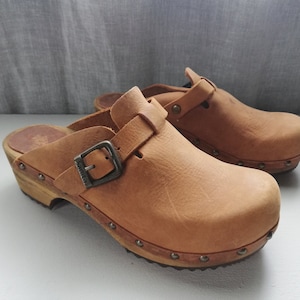 May include: A pair of brown leather clogs with a buckle strap. The clogs have a wooden sole with metal studs.