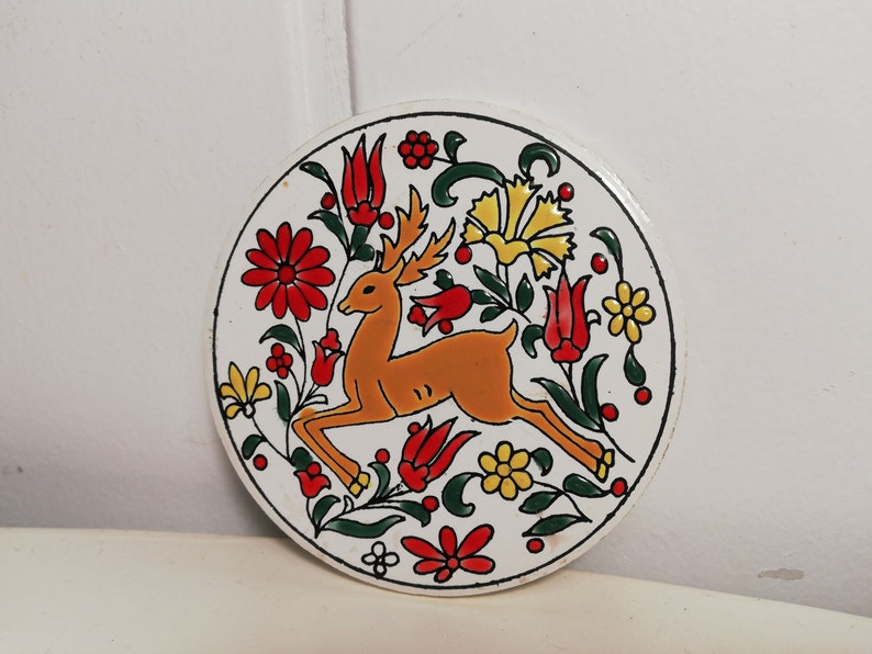 Puede incluir: Un plato redondo de cer&aacute;mica blanca con un dise&ntilde;o pintado a mano de un ciervo naranja y marr&oacute;n, rodeado de flores rojas, amarillas y verdes.