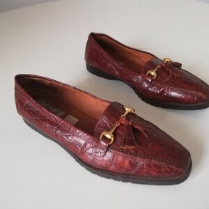 Puede incluir: Un par de mocasines de cuero burdeos con detalles dorados y borlas. Los mocasines tienen un patr&oacute;n de piel de cocodrilo.