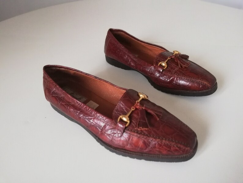 Puede incluir: Un par de mocasines de cuero burdeos con detalles dorados y borlas. Los mocasines tienen un patr&oacute;n de piel de cocodrilo.