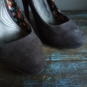 Op de afbeelding: Een paar zwarte su&egrave;de pumps met hoge hakken en een bloemenprint voering.